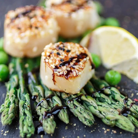 Three seared scallops, topped with Gustus Vitae Essential Spices from the Pantry Starter Kit, are drizzled with sauce and served on asparagus, accented by green peas and lemon wedges in the background. Brand: vendor-unknown.