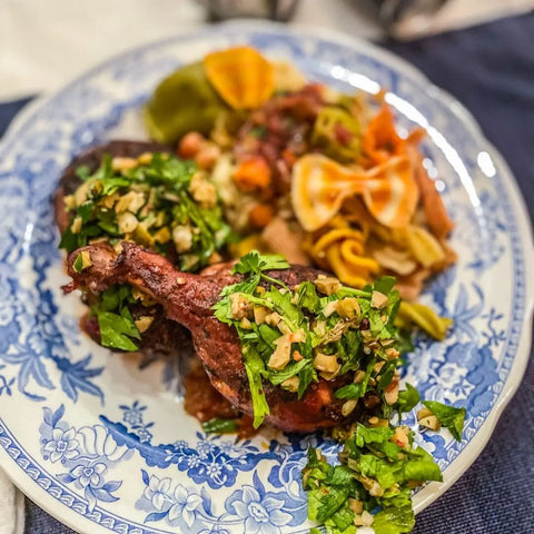A blue and white plate with roasted chicken legs topped with herbs and nuts using spices from the Pantry Starter Kit (8 Magnetic Tins by vendor-unknown), served with colorful veggies and pasta.