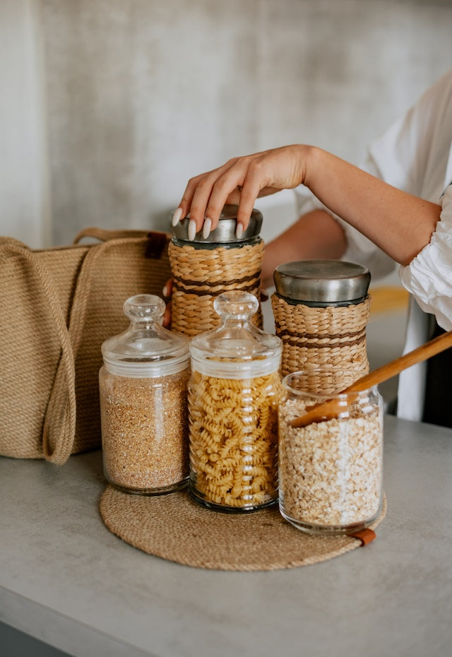 Person reaching for a jar of food on a table with various jars and a woven bag.