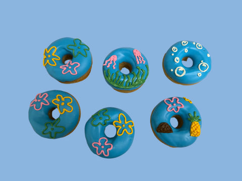 Dogtown Bakery’s Sponge Bob Doughmutts features six donut-shaped gourmet dog treats with blue icing and ocean-themed designs—starfish, jellyfish, bubbles, tropical flowers, pineapple, and seashell—arranged in two rows.