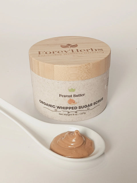 A jar of Forev'Herbs Peanut Butter Organic Whipped Sugar Scrub rests on a white surface, with a white spoon in front holding a dollop of the shea butter-infused exfoliant, which looks just like creamy peanut butter.