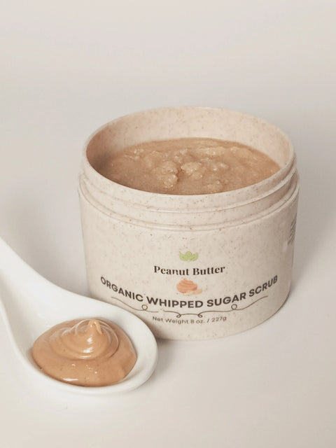 An open jar of Forev'Herbs Peanut Butter Organic Whipped Sugar Scrub sits on a white surface beside a white ceramic spoon holding some of the shea butter exfoliant.