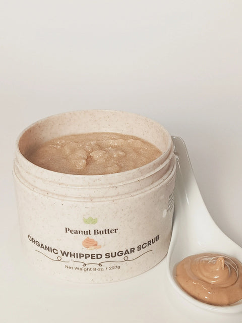 An open jar of Forev'Herbs Peanut Butter Organic Whipped Sugar Scrub sits beside a white spoon with a dollop of the light brown exfoliator, resembling creamy peanut butter—a luxurious natural body scrub.