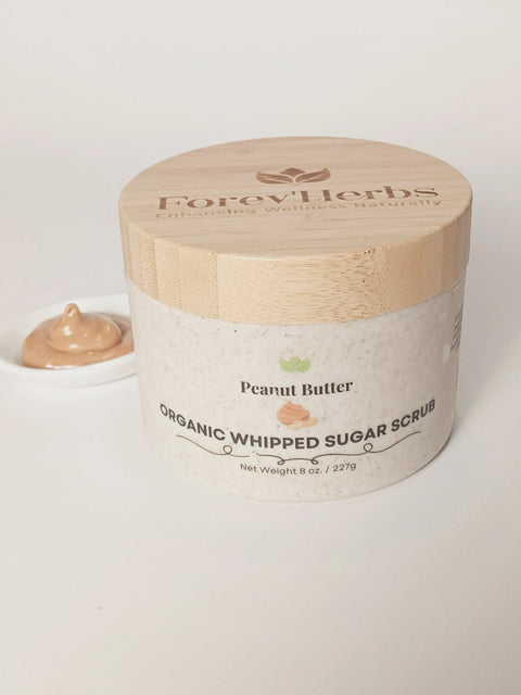 A jar of Forev'Herbs Peanut Butter Organic Whipped Sugar Scrub with a wooden lid is placed next to a small dish with a dollop of the shea butter exfoliant, all against a white background.