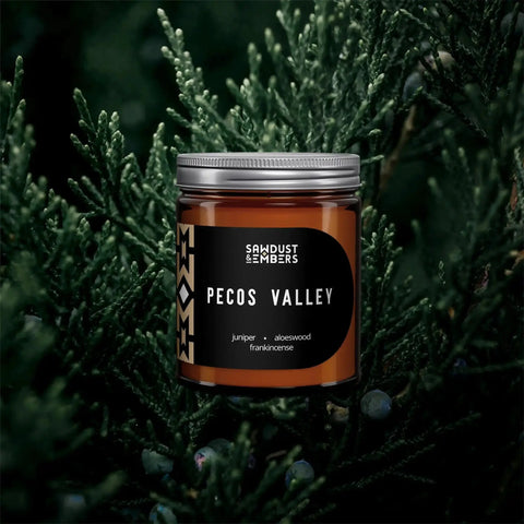 A brown Pecos Valley - Signature Candle by Sawdust & Embers rests amid green juniper branches, its label highlighting notes of juniper, aloeswood, and frankincense—a tribute to artisanal home fragrances.