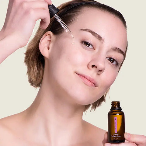 A person with short hair uses a dropper to apply Open Formula Collagen Peptide Serum from a brown glass bottle to their cheek, smiling gently and highlighting smooth skin against a neutral beige background.