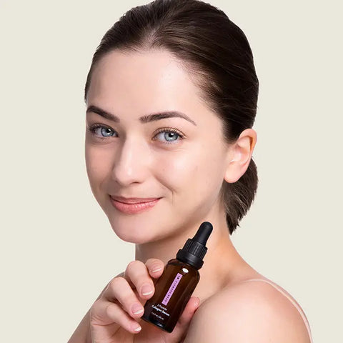 A young woman with fair skin and dark hair pulled back smiles softly while holding a brown glass dropper bottle of Open Formula Collagen Peptide Serum, suggesting its use to enhance skin texture.