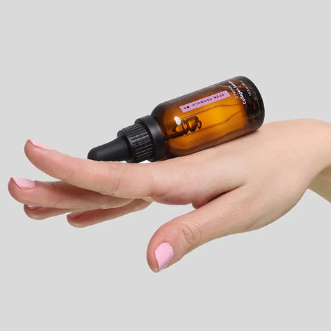 A hand with light pink nails balances an Open Formula Collagen Peptide Serum brown glass dropper bottle on the back of the fingers against a plain light gray background.