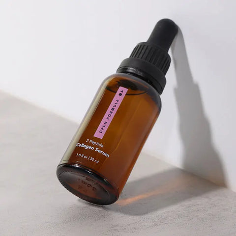 A brown glass dropper bottle labeled Collagen Peptide Serum by Open Formula, featuring a pink stripe, rests at an angle on a white surface—hinting at smoother skin texture from collagen peptides.