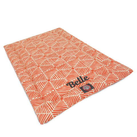 The Majestic Pet Charlie Crate Mat - Personalized is a rectangular orange dog bed with a geometric pattern, the name Belle, and paw print logo in one corner. Its machine washable, soft, cushioned, elevated, and ideal as a crate mattress.