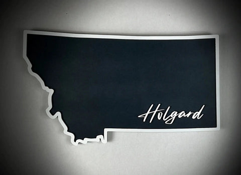 The Medium Floating MT Outline Sign Personalized by B. James Design & Woodwork features a dark Montana silhouette with Holgard in white cursive at the bottom right—ideal custom home décor on your choice of background color.
