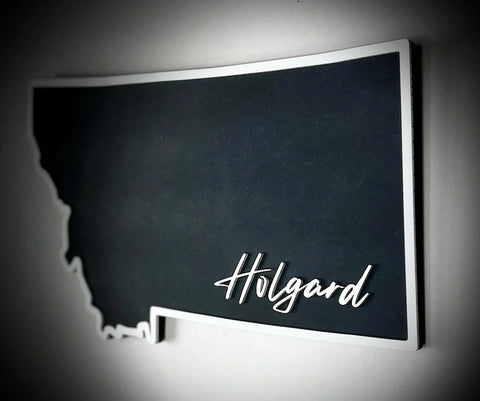 The Medium Floating MT Outline Sign Personalized by B. James Design & Woodwork is a Montana-shaped wall sign with a dark background and Holgard in white script at the bottom right, perfect for home decor.
