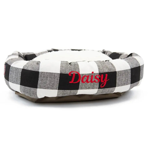 The Majestic Pet Anderson Check Sherpa Bagel Bed - Personalized features a round black and white plaid design, raised edges for spine support, a soft cushioned interior, and red embroidered name “Daisy” on the side.
