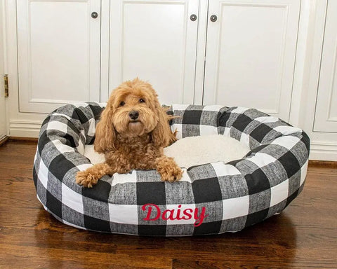 A fluffy light brown dog sits in the Majestic Pet Anderson Check Sherpa Bagel Bed - Personalized, with “Daisy” embroidered in red on the front, offering excellent spine support on a wooden floor by white cabinets.