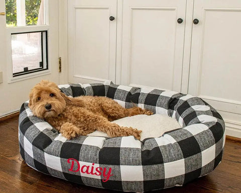 A fluffy, light brown dog lounges in a personalized Anderson Check Sherpa Bagel Bed by Majestic Pet, featuring Daisy in red letters, set in a room with white cabinets and a wooden floor.
