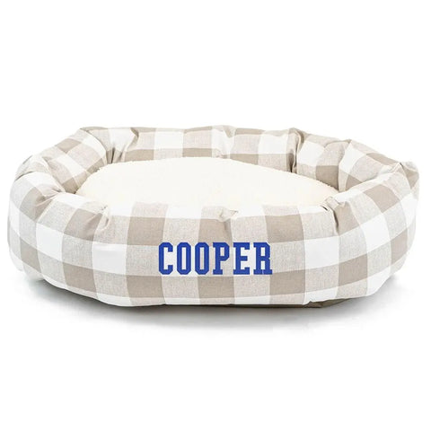 The Anderson Check Sherpa Bagel Bed by Majestic Pet features a soft, cushioned center for spine support, a beige and white checkered design, and can be personalized with your pets name in bold blue letters on the front.
