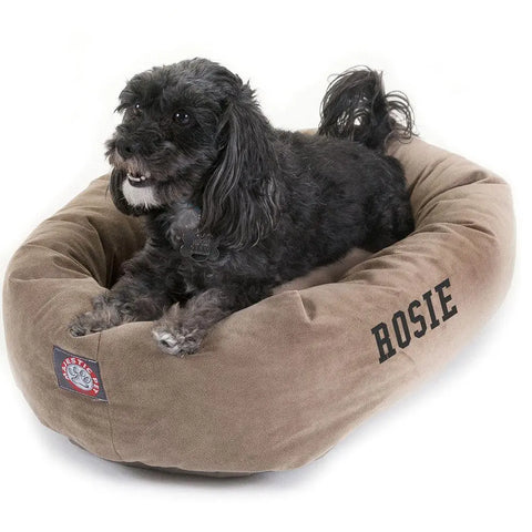 A small black dog with curly fur rests on a tan Majestic Pet Suede Bagel Dog Bed—Personalized with ROSIE. The cozy bed features a sewn patch and removable slipcover, keeping the alert pup comfortable as it lounges.