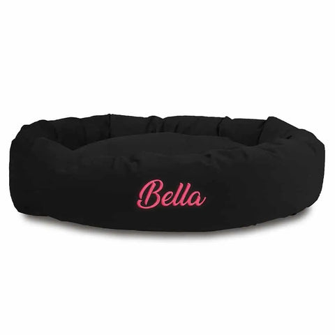 The Majestic Pet Suede Bagel Dog Bed is round and black, with Bella embroidered in pink cursive on the front. It has a removable slipcover for easy cleaning and can be personalized.