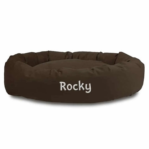 The Majestic Pet Suede Bagel Dog Bed - Personalized is a round, brown suede bed with soft, raised edges and Rocky embroidered in white. It features a plush design and a removable slipcover for easy cleaning and lasting comfort.