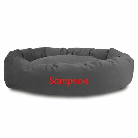 The Majestic Pet Suede Bagel Dog Bed features a round, gray suede design, a removable slipcover, water-resistant fabric for easy care, and “Sampson” embroidered in red on the front. Personalize it for your dog!.