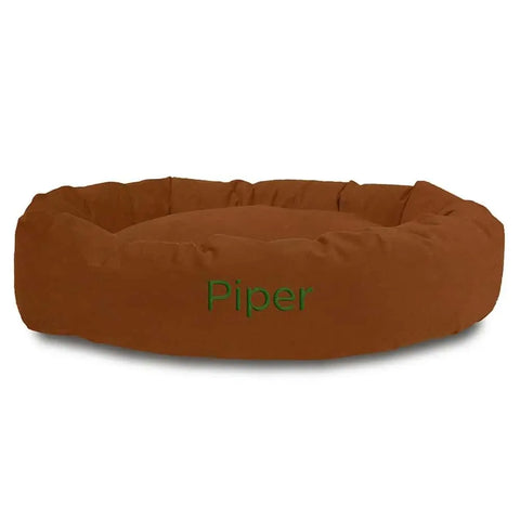 The Majestic Pet Suede Bagel Dog Bed features a round, brown suede design with a water-resistant, removable slipcover and Piper embroidered in green on the front.