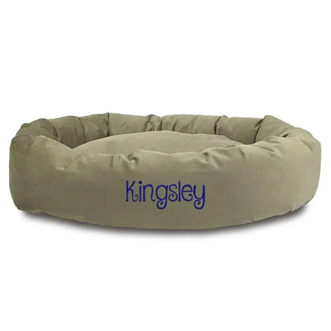The Majestic Pet Suede Bagel Dog Bed features light brown suede, raised edges, a plush center, and a removable slipcover. Personalize it with your dogs name, embroidered in blue on the front.