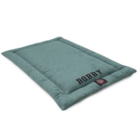 The Villa Crate Mat - Personalized by Majestic Pet features a rectangular, soft teal design with raised edges. Bobby and a cartoon dog patch are embroidered on one side. Plush, comfy, and machine washable for easy care.