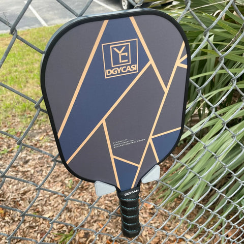 A DGYCASI pickleball paddle with a gold and blue geometric design hangs on a fence using the Chatelet Manufacturing Pickleball Paddle Chain Link Fence Holder—compact, secure, USA-made—near green plants and a parking lot.