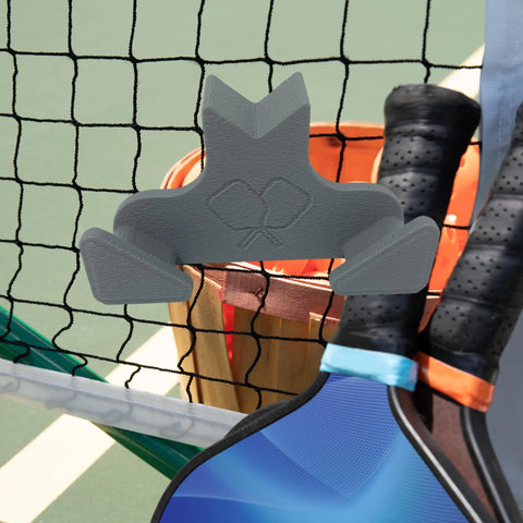 The Chatelet Manufacturing Pickleball Paddle Chain Link Fence Holder, a compact USA-made accessory, securely mounts your paddle on the court; shown hanging on a net beside two paddles with a wooden basket and green court in the background.