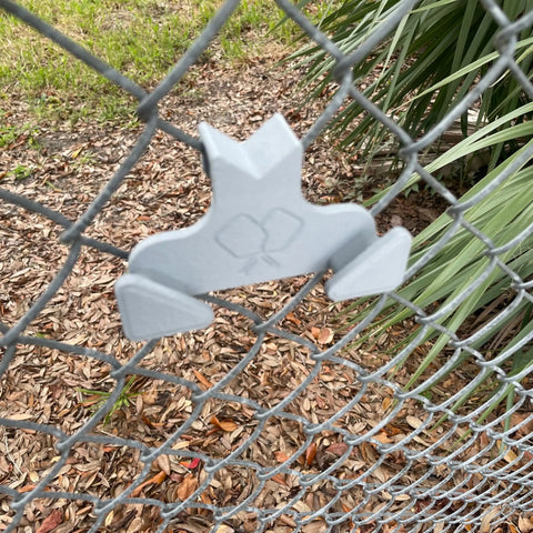 The Chatelet Manufacturing Pickleball Paddle Chain Link Fence Holder, a gray metal frog-shaped accessory, securely mounts paddles to the court fence; mulch, grass, and green plants appear in the background.