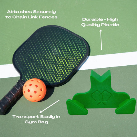 A pickleball paddle and orange ball sit on a court next to the Chatelet Manufacturing Pickleball Paddle Chain Link Fence Holder, which attaches securely to fences, is made of durable plastic, and transports easily in any gym bag.