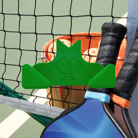 The Chatelet Manufacturing Pickleball Paddle Chain Link Fence Holder secures your paddle on the court net. Compact and made in the USA, its shown here holding a paddle, with balls, paddles, and a basket visible on the court.