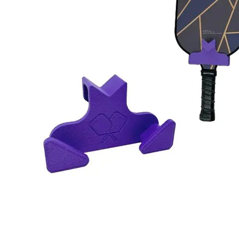 A purple Chatelet Manufacturing Pickleball Paddle Chain Link Fence Holder, featuring an engraved paddle and ball design, is shown attached next to a pickleball paddle handle.