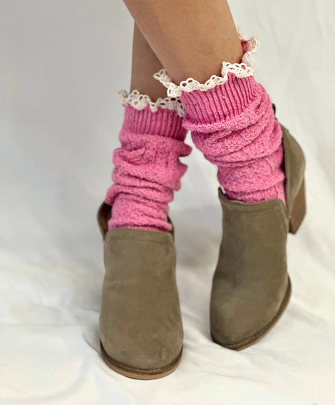 Close-up of crossed legs wearing dusty brown slip-on shoes and Catherine Cole BOOTIE Lace Slouch Socks in pink with ruffled trim, set against a plain white background.