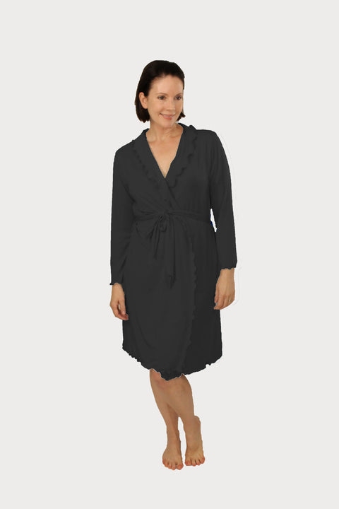 A woman with short dark hair stands barefoot, smiling in the knee-length black Goodnighties Lettuce-Edge Robe featuring long sleeves, a tie waist, and ruffled edging, set against a plain white background.