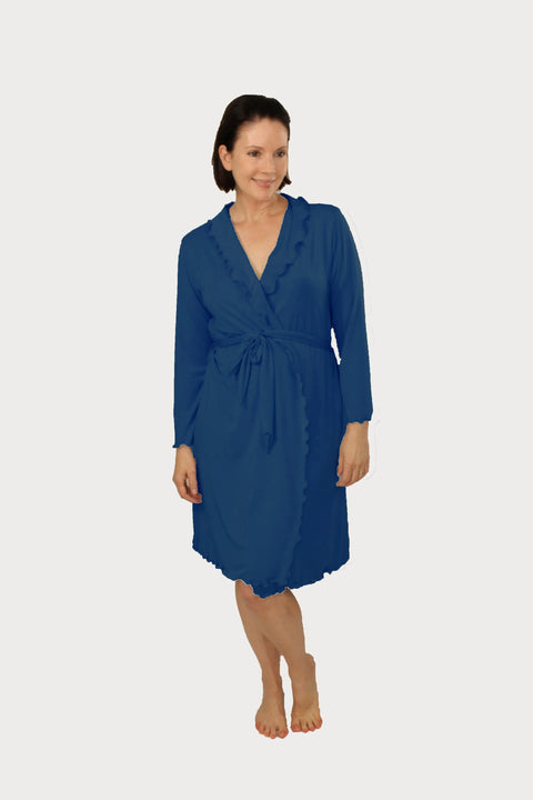 A woman with short brown hair stands barefoot, smiling and looking slightly right, wearing the Goodnighties Lettuce-Edge Robe in blue—an American-made, knee-length robe with a waist tie and ruffled edges—against a white background.