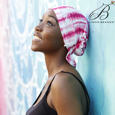 A smiling woman in a pink and white Tanisha Chemo Beanies® scarf beanie leans against a blue patterned wall, looking upward. The Chemo Beanies® logo appears in the top right corner.