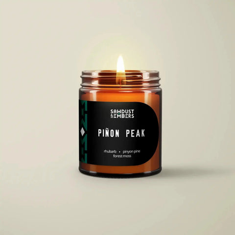 A lit Sawdust & Embers Piñon Peak - Signature Candle, featuring notes of rhubarb, pinon pine, and forest moss, sits on a light beige surface—an inviting handcrafted soy candle ideal for adding mountain-inspired fragrance.