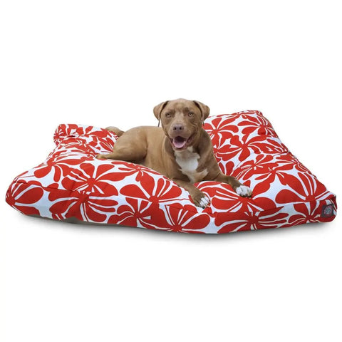 A brown dog with white markings lies on a Majestic Pet Plantation Rectangle Dog Bed with premium polyester fill, featuring a large red and white floral pattern, looking at the camera with its mouth open and tongue out.