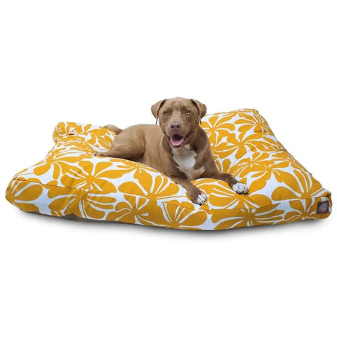 A brown dog with a white chest lies on a large Majestic Pet Plantation Rectangle Dog Bed with a yellow and white floral pattern, looking at the camera with its mouth open.