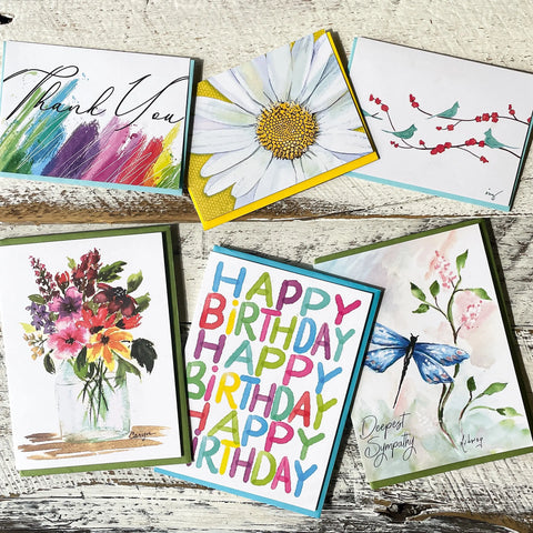 The NEW Assorted Card Bundle from JuLu Cards features six colorful designs, including Thank You, daisy, cherry blossoms, floral bouquet, Happy Birthday, and butterfly with Deepest Sympathy on a wooden surface.