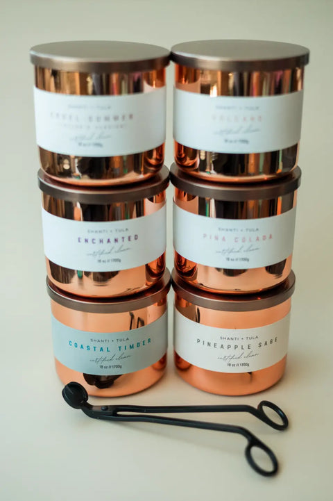 Six copper-colored candles with white labels, in scents like Coffee Vanilla and Pineapple Sage, are stacked in two columns. A Shanti + Tula Premium Candle Wick Trimmer, handcrafted in the USA to improve burn quality, sits beside them.