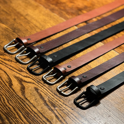 Six Baxter Leather Belts by Rogue Industries, each 1.5 wide and crafted from premium cowhide leather in brown and black, are arranged in parallel on a wooden surface, with their metal buckles aligned at the bottom left of the image.