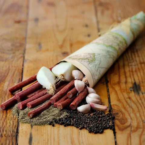 A Field Trip Gift Cooler by Field Trip displays cured meat sticks, a halved onion, garlic cloves, ground black pepper, and whole peppercorns on a rustic wooden table.