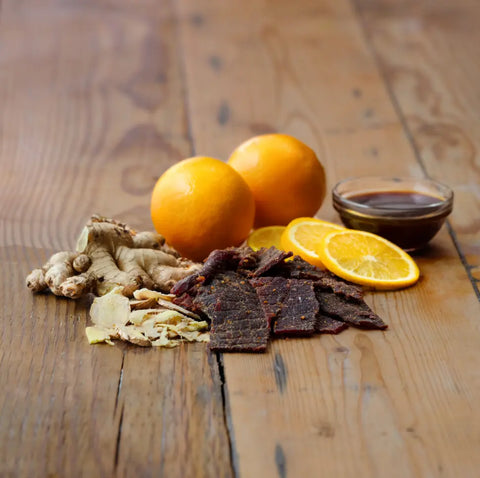 Beef jerky slices, fresh ginger, and whole oranges are arranged on a rustic table—perfect for the Field Trip Gift Cooler by Field Trip or as part of a unique jerky gift set.