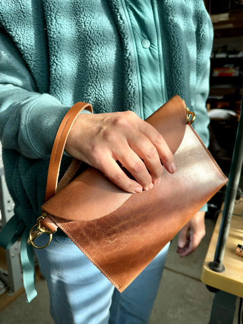 A person in a teal fleece jacket and light blue pants holds the Homestead Brand Standard Wristlet No. 1604, a full grain leather clutch with a curved flap and gold hardware, indoors—possibly in a workshop.