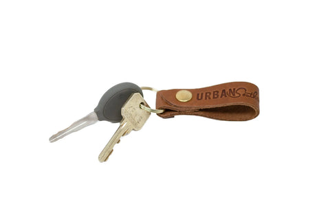 Two keys on a metal keyring are attached to the American-made Urban Key Fob by Urban Southern, crafted from genuine leather with URBAN soul stamped and a brass button. The background is white.
