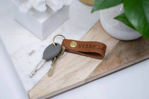 A set of keys with an Urban Southern Urban Key Fob made from genuine American leather rests on a wooden and marble surface near a white decorative item and a green plant.