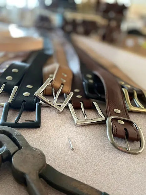 A close-up of BBH Handcrafted Leather Belts in black and brown with metal buckles lies on a table, accompanied by a metal tool and a small nail in the foreground, with a blurred background.