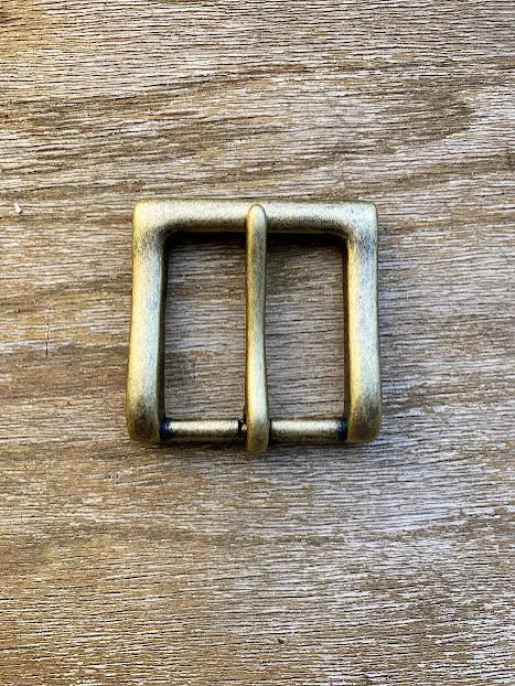 A brass rectangular belt buckle with a central prong lies flat on textured wood—an ideal match for the BBH Handcrafted Leather Belt, highlighting artisan quality and style.